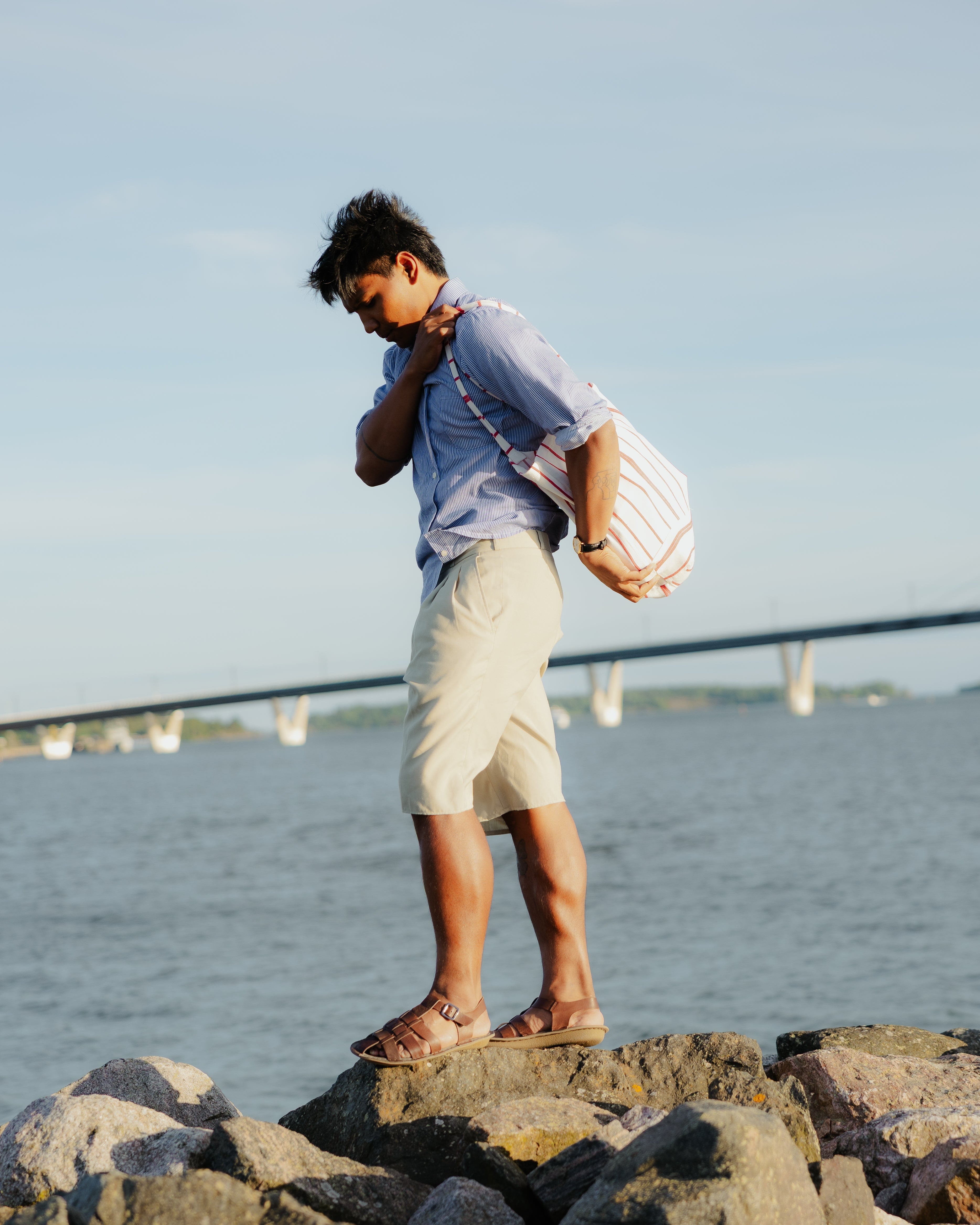 Man wearing cocoa brown leather shoulder bag with cream pants and blue shirt, standing on rocks by water.